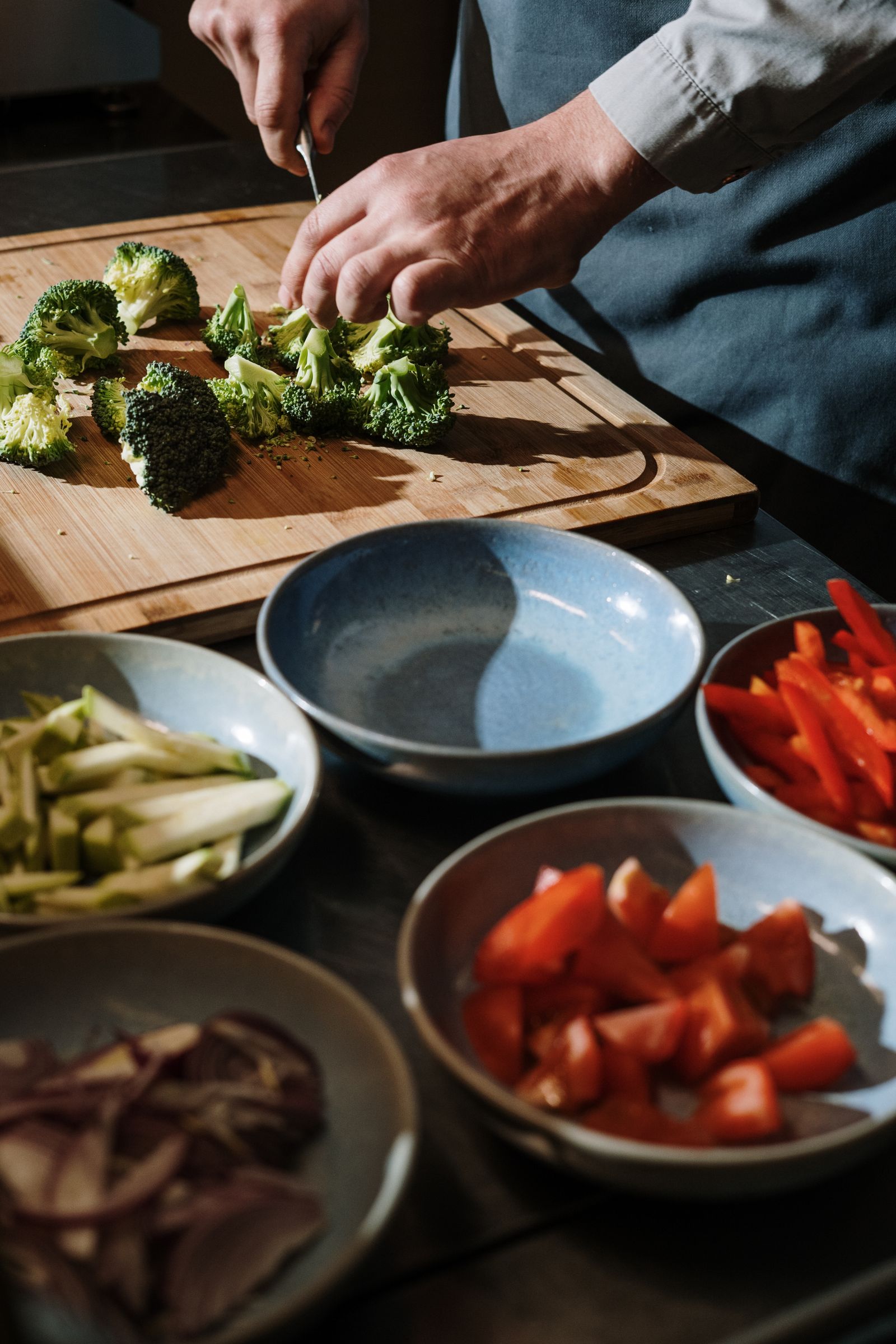 Hands chopping fresh broccoli on a wooden board with bowls of prepared peppers, tomatoes and onions
