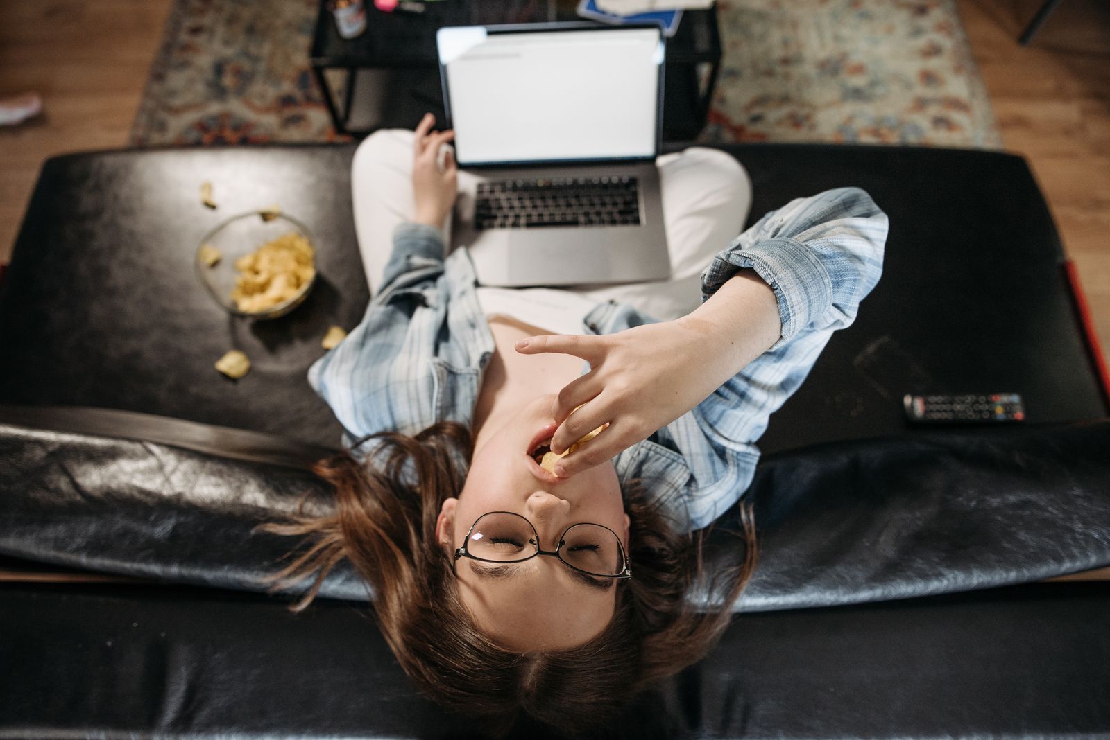 Overhead view of a woman eating crisps from a bowl while working at a laptop on a sofa